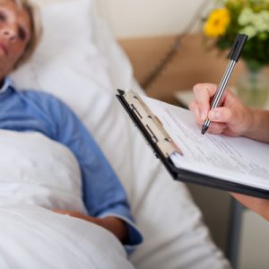 A white woman in a blue gown lying in a hospital bed in the background, with someone holding a clipboard and pen in the foreground. The woman may be receiving uncompensated care.