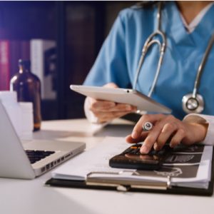 Hospital doctor sits at a desk with a laptop, clipboard, and phone.